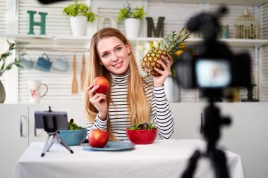 Yemek blogcusu mutfak stüdyosunda taze meyve salatası pişiriyor video kanalı için özel ders kameraları önünde çekim yapıyor. Kadın nüfuzlu kişi elmayı, ananası tutuyor ve sağlıklı beslenmekten bahsediyor. Fructorianism