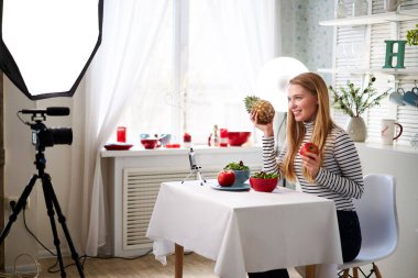 Yemek blogcusu mutfak stüdyosunda taze meyve salatası pişiriyor video kanalı için özel ders kameraları önünde çekim yapıyor. Kadın nüfuzlu kişi elmayı, ananası tutuyor ve sağlıklı beslenmekten bahsediyor. Fructorianism