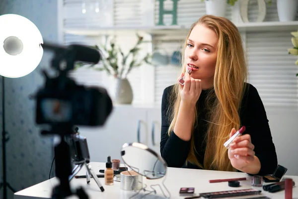 Beauty blogger woman filming daily make-up routine tutorial near camera on tripod. Influencer blonde girl live streaming cosmetics product review in home studio. Vlogger female comparing lipsticks. — Stock Photo, Image