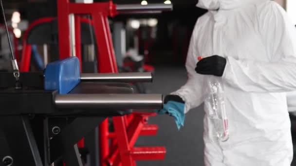 Hazmat pracovník dezinfikuje tělocvičnu fitness vybavení z coronavirus covid-19 nebezpečí s antibakteriální dezinfekční rozprašovač a otřít. Muž v ochranném obleku čistí výcvikový přístroj na cvičišti. — Stock video