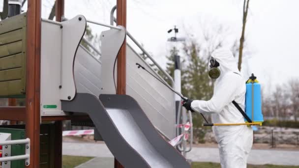 Hazmat team disinfects playground surfaces on coronavirus covid-19 quarantine with antibacterial sanitizer sprayer. Worker in gas mask and protective suit decontaminates children rides, slide, street. — Stock Video