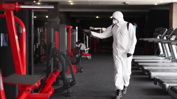 Hazmat arbetare desinficerar gym träningsutrustning från coronavirus covid-19 fara med antibakteriella rengöringsmedel spruta på karantän. Man i skyddsdräkt rengör träningsredskap på träningsområdet. — Stockvideo