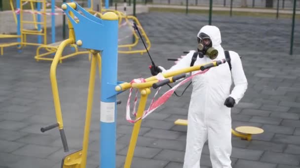 Hazmat werknemer desinfecteert sportvelden oppervlakken van coronavirus covid-19 gevaar met antibacteriële ontsmettingsspray op quarantaine. Man in beschermend pak reinigt openbare atletische fitnesstoestellen. — Stockvideo