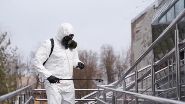 El desinfectante desinfecta las superficies táctiles de la barandilla en la cuarentena del coronavirus covid-19 con pulverizador antibacteriano. Trabajador en máscara de gas y traje protector de materiales peligrosos limpia pasamanos en el lugar público de la ciudad . — Vídeos de Stock