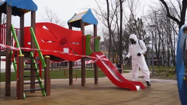Hazmat team disinfects playground surfaces on coronavirus covid-19 quarantine with antibacterial sanitizer sprayer. Worker in gas mask and protective suit decontaminates children rides, slide, street. — Stock Video