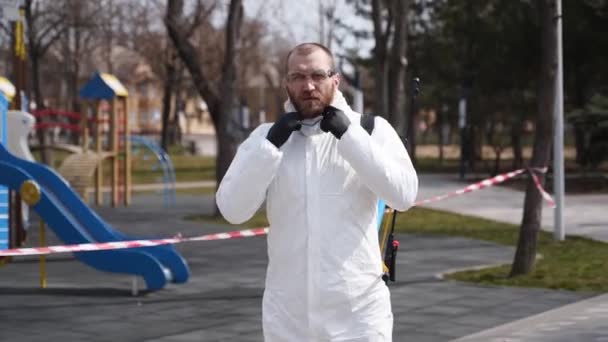 Gevaarlijke materialen verwijderen werknemer draagt beschermende hazmat pak en gezichtsmasker met sanitaire voorzieningen poseren tijdens virusuitbraak. Heldenschot van een man in een ademhalingsapparaat met sproeier. Coronavirus, COVID-19 — Stockvideo