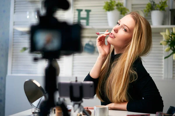 Vlogger fêmea aplica batom nos lábios. Mulher blogueira de beleza filmando tutorial de rotina de maquiagem diária na câmera no tripé. Influenciador menina loira ao vivo streaming cosméticos revisão do produto em estúdio em casa — Fotografia de Stock