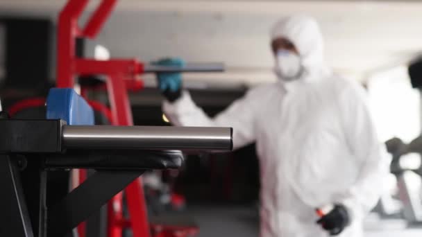 Hazmat worker disinfects gym fitness equipment from coronavirus covid-19 hazard with antibacterial sanitizer sprayer and wipe. Man in protective suit cleans training apparatus at workout area. — Stock Video