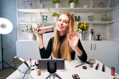 Güzellik blogcusu kadın tripodla kameranın yanında günlük makyaj rutin dersini çekiyor. Etkilenen kız canlı yayın kozmetik ürünleri eleştirisi ev stüdyosunda. Vlogger dişisi göz farı paleti gösteriyor.