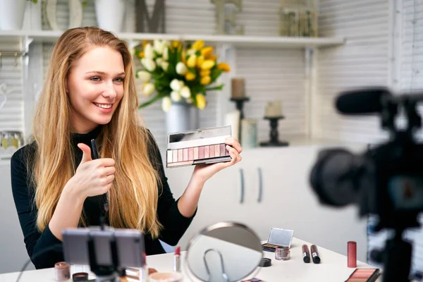 Beauty blogger woman filming daily make-up routine tutorial near camera. Influencer girl live streaming cosmetics product review. Vlogger female recommends eye shadow palette showing thump up sign. — Stock Photo, Image