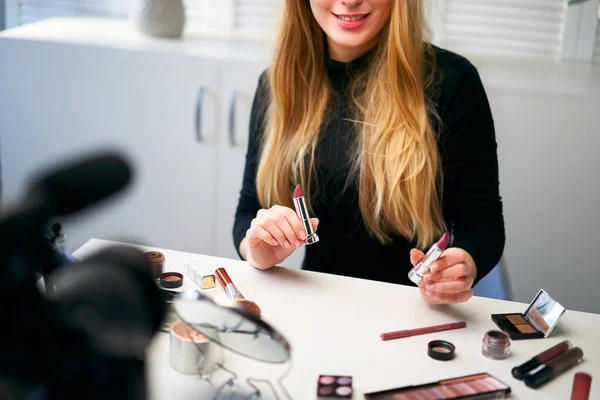 Beauty blogger woman filming daily make-up routine tutorial near camera on tripod. Influencer blonde girl live streaming cosmetics product review in home studio. Vlogger female comparing lipsticks. — Stock Photo, Image