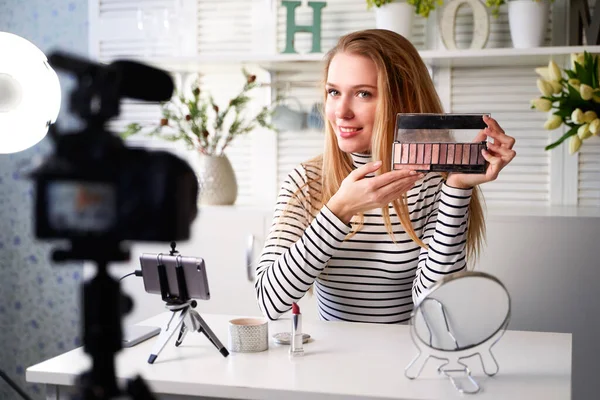 Beauty blogger woman filming daily make-up routine tutorial near camera on tripod. Influencer girl live streaming cosmetics product review in home studio. Vlogger female showing eye shadow palette. — Stock Photo, Image