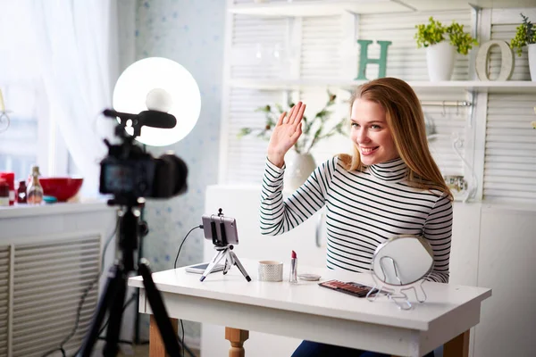 Female beauty blogger in white outfit greets fans while sitting in front of camera and waving her hand. Influencer woman filming video blog and reviews cosmetics. Make-up artist recording vlog. — Stock Photo, Image