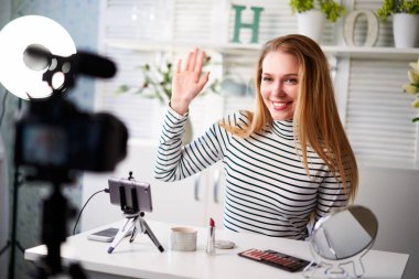 Beyaz kıyafetli kadın güzellik blogcusu, kameranın önünde oturup elini sallarken hayranlarını selamlıyor. Nüfuzlu kadın video blogu çekiyor ve kozmetik eleştirileri yapıyor. Makyaj sanatçısı vlog kaydediyor.