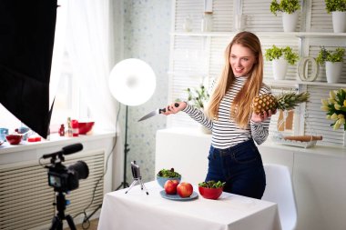 Yemek blogcusu mutfak stüdyosunda taze meyve salatası pişiriyor video kanalı için özel ders kameraları önünde çekim yapıyor. Kadın nüfuzlu kişi elmayı, ananası tutuyor ve sağlıklı beslenmekten bahsediyor. Fructorianism