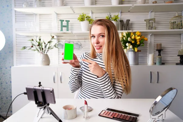 Beauty blogger woman filming advertising, holding green screen smartphone application. Make-up influencer blonde girl live streaming cosmetics product review in studio asking for likes, subscription