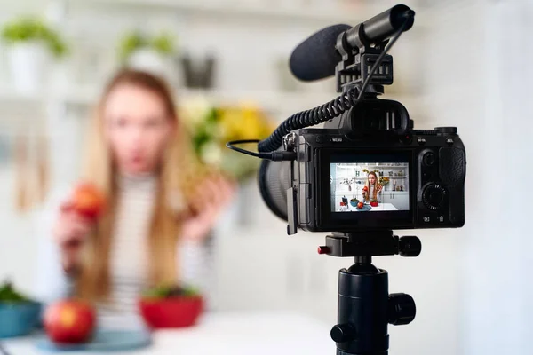 Display of camera recording video blog for food blogger woman with apple, pineapple in kitchen studio talking about healthy vegan eating. Influencer vlogger girl live streaming nutrition masterclass.