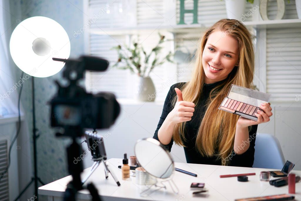 Beauty blogger woman filming daily make-up routine tutorial near camera. Influencer girl live streaming cosmetics product review. Vlogger female recommends eye shadow palette showing thump up sign.