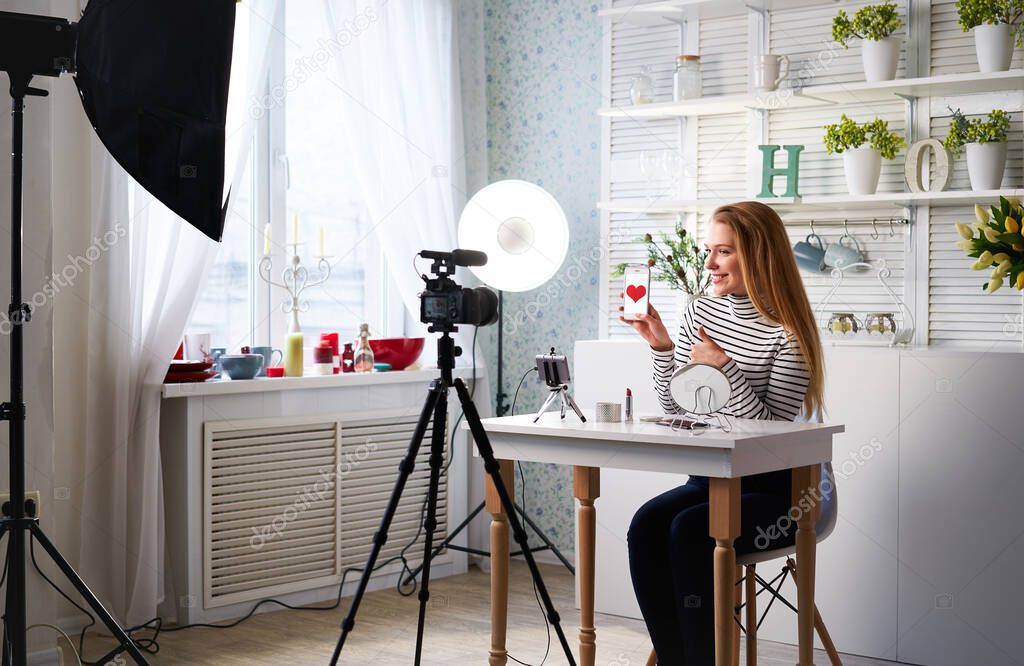 Beauty blogger woman filming make-up asks for likes, thumbs up, subscription on camera. Influencer blonde girl live streaming cosmetics product review in studio with professional lighting equipment.