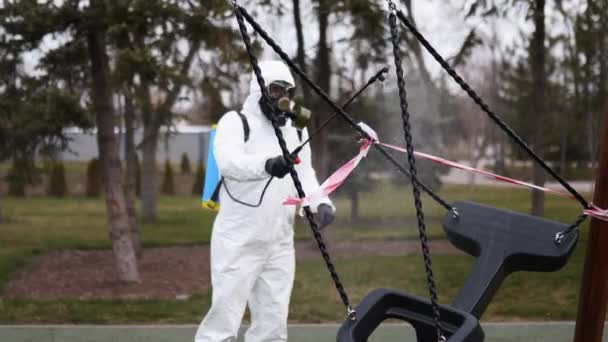Hazmat worker disinfects playground surfaces with antibacterial sanitizer sprayer on coronavirus covid-19 quarantine. Man in gas mask and protective suit decontaminates children swings in public area. — Stock Video