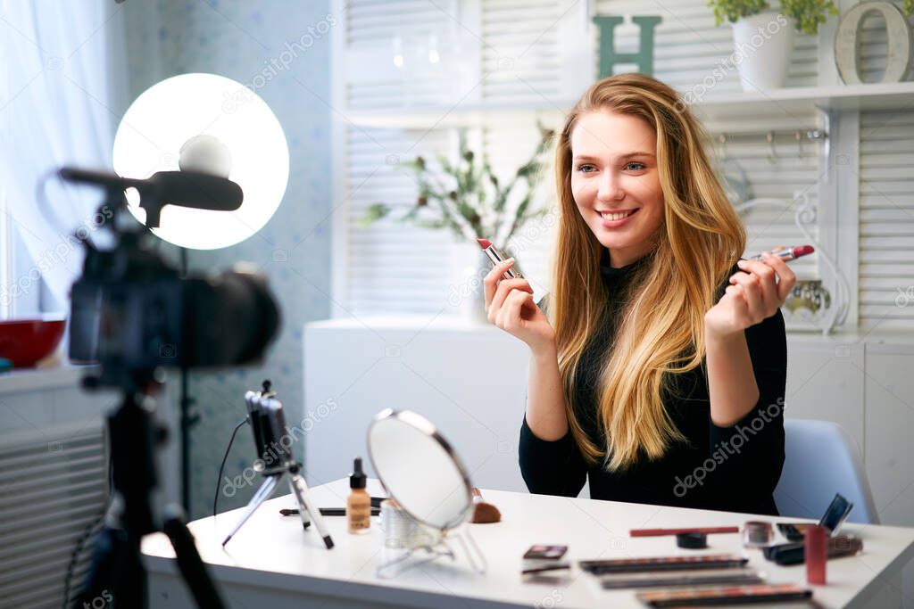 Beauty blogger woman filming daily make-up routine tutorial near camera on tripod. Influencer blonde girl live streaming cosmetics product review in home studio. Vlogger female comparing lipsticks.