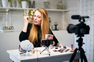 Güzellik blogcusu kadın tripodla kameraya günlük makyaj dersini çekiyor. Etkilenen kız canlı yayın kozmetik ürünleri eleştirisi ev stüdyosunda. Vlogger dişisi fırçayla cilt pudrası uyguluyor.
