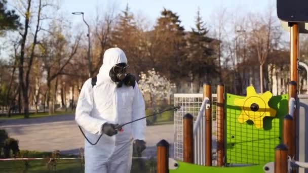 Hazmatチームは、コロナウイルスcovid-19隔離の遊び場表面を抗菌消毒剤噴霧器で消毒します。ガスマスクと保護スーツの労働者は子供の乗り物、スライド、通りを除染. — ストック動画