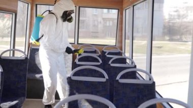 Tehlikeli madde ekibi çalışanı otobüsün içini antibakteriyel dezenfektanla dezenfekte ediyor. Coronavirüs covid-19 karantinasında. Gaz maskeli adam, tehlikeli madde giysisi toplu taşıma koltuklarını temizliyor, bez parçasıyla tutunuyor..