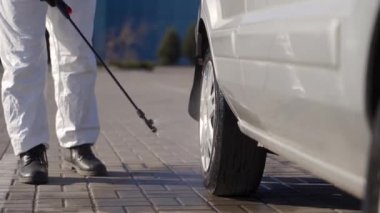 Tehlikeli madde ekibi çalışanı araba tekerleklerini antibakteriyel dezenfektanla dezenfekte ediyor. Coronavirüs covid-19 karantinasına. Gaz maskeli adam, tehlikeli madde giysisi park etmiş arabanın lastiklerini ve vücudunu basınç makinesiyle temizliyor..