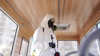 Koronavirüs covid-19 karantinasında antibakteriyel dezenfektan spreyli tehlikeli madde ekibi çalışanı. Gaz maskeli bir adam toplu taşıma araçlarını çamaşır makinesiyle dezenfekte ediyor ve kameraya bakıyor..