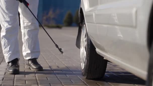 Hazmat team worker disinfects car wheels with antibacterial sanitizer sprayer on coronavirus covid-19 quarantine. Man in gas mask, hazmat suit cleans parked car tires and body with pressure washer. — Stock Video