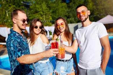 Bir grup arkadaş, havuz başında yaz partisinde bardakları tokuşturup güneşli bir günde havuz kenarında kokteyl içiyorlar. İnsanlar tropikal tatillerde lüks villada taze meyve suyu içerek kadeh kaldırıyorlar..