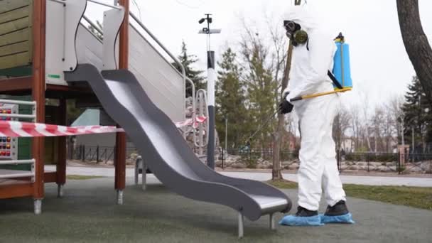 Hazmat team disinfetta le superfici del parco giochi su coronavirus covid-19 quarantena con spruzzatore disinfettante antibatterico. Operaio in maschera antigas e tuta protettiva decontamina bambini giostre, scivolo, strada . — Video Stock