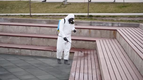 Operatore di rimozione materiali pericolosi disinfetta banchi con spruzzatore disinfettante antibatterico su coronavirus covid-19 quarantena. Uomo in maschera antigas, tuta hazmat pulisce pubblico stabilirsi sedie nel parco della città  . — Video Stock