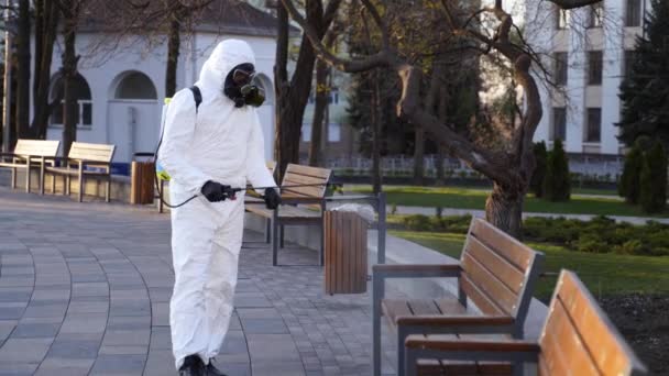 Hazmat team medewerker desinfecteert banken met antibacteriële ontsmettingsspray op coronavirus covid-19 quarantaine. Man met gasmasker, hazmat pak reinigt openbare stoelen in het park met drukreiniger. — Stockvideo