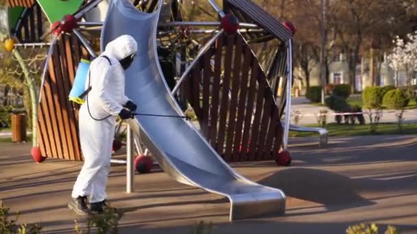 Hazmat team disinfects playground surfaces on coronavirus covid-19 quarantine with antibacterial sanitizer sprayer. Worker in gas mask and protective suit decontaminates children rides, slide, street. — Stock Video