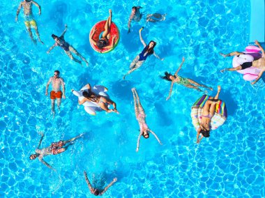 Havuzda, şişme flamingo, kuğu ve şiltelerle parti veren arkadaşlar. Mutlu gençler güneşli bir günde lüks bir tatil köyünde dinleniyorlar. Yukarıdan bak. Bikinili kızlar güneşte güneşleniyor..