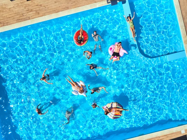 Aérea de amigos teniendo fiesta en piscina con flamenco inflable, cisne, colchón. Los jóvenes felices saludan con las manos a la cámara de drones en un resort de lujo. Vista desde arriba. Chicas en bikini tomando el sol. —  Fotos de Stock