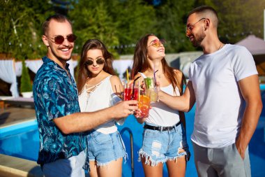 Bir grup arkadaş, havuz başında yaz partisinde bardakları tokuşturup güneşli bir günde havuz kenarında kokteyl içiyorlar. İnsanlar tropikal tatillerde lüks villada taze meyve suyu içerek kadeh kaldırıyorlar..