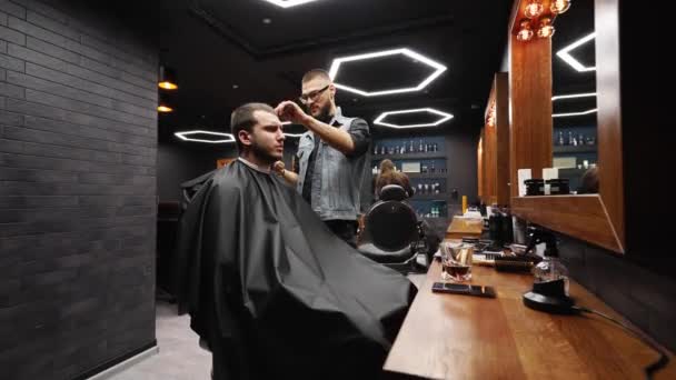 Trendig frisör klipper skäggiga män hår med en klippare i frisörsalongen. Män frisyr och hårklippning i salong. Grooming håret med trimmer. Hår frisör gör frisyr i retro frisör. Dolly in — Stockvideo