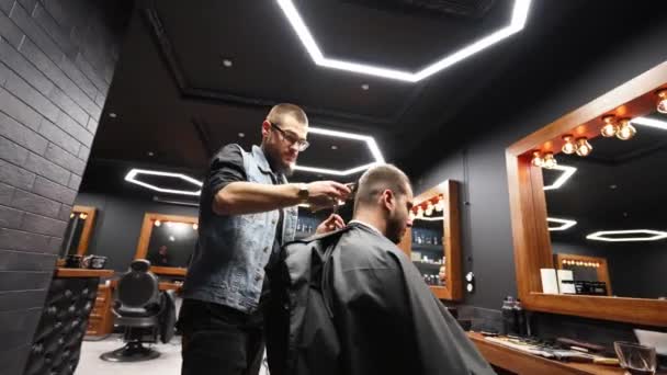 Trendy barber cuts bearded mans hair with a clipper in barbershop. Mens hairstyling and hair cutting in salon. Grooming the hair with trimmer. Hairdresser doing haircut in retro hair salon. Tracking — Stock Video