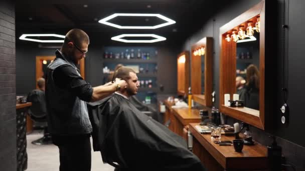 Trendy barber cuts bearded mans hair with a clipper in barbershop. Mens hairstyling and hair cutting in salon. Grooming the hair with trimmer. Hairdresser doing haircut in retro hair salon. Tracking — Stock Video