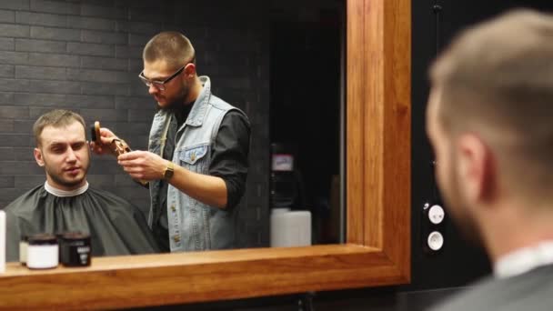 Barber in glasses cuts bearded mans hair with a clipper in barbershop. Hipster hairstyling and hair cutting in salon. Grooming hair with trimmer. Hairdresser doing haircut in hair salon. Mirror shot. — Stock Video