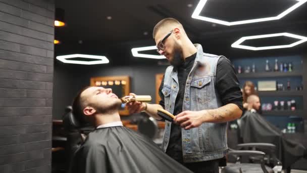 Frisör i glasögon skär mans skägg med en trimmer i frisörsalongen. Hårstyling och hårklippning i salong. Grooming håret med clipper. Hipster frisör gör frisyr i retro frisör. — Stockvideo