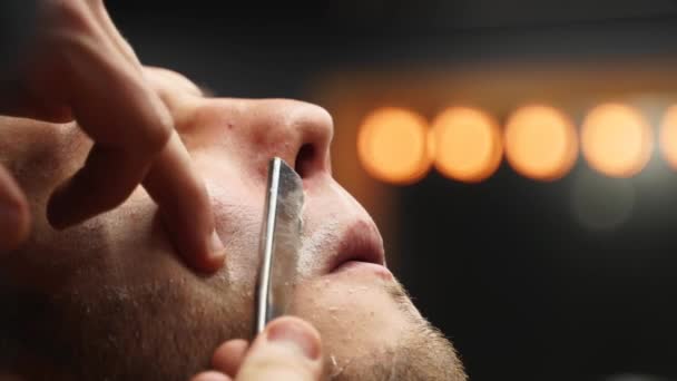 Professional barber shaves customer beard with straight razor. Beard cut with old-fashioned blade at barbershop. Handsome macho man getting his beard shaved in studio. Close-up shot. — Stock Video