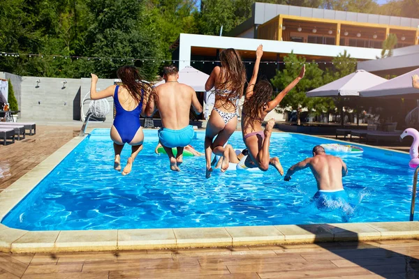 Amigos felizes pulando e salpicando na piscina com flutuadores infláveis em resort de luxo. Jovens em roupa de banho fazendo festa em casa de férias privada no dia ensolarado. Meninas quentes pular na água. — Fotografia de Stock