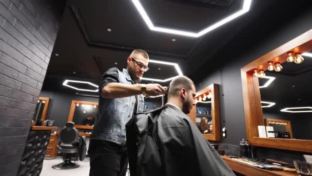 El peluquero moderno corta el cabello de los hombres barbudos con un cortador en la barbería. Para hombre peinado y corte de pelo en el salón. Aseo el cabello con trimmer. Peluquería haciendo corte de pelo en peluquería retro. Seguimiento — Vídeo de stock
