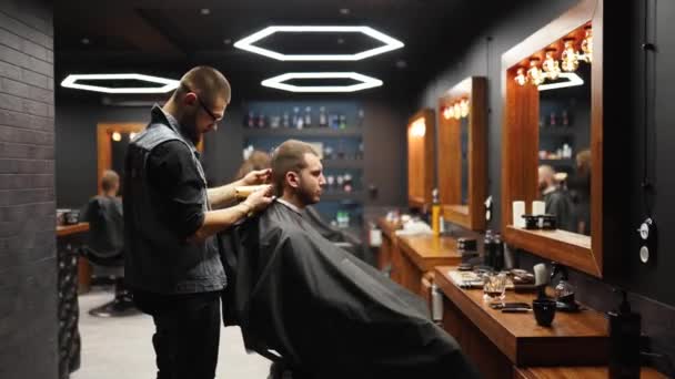Trendy barber cuts bearded mans hair with a clipper in barbershop. Mens hairstyling and hair cutting in salon. Grooming the hair with trimmer. Hairdresser doing haircut in retro hair salon. Tracking — Stock Video