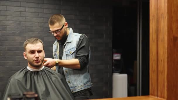 Barber in glasses cuts bearded mans hair with a clipper in barbershop. Hipster hairstyling and hair cutting in salon. Grooming hair with trimmer. Hairdresser doing haircut in hair salon. Mirror shot. — Stock Video