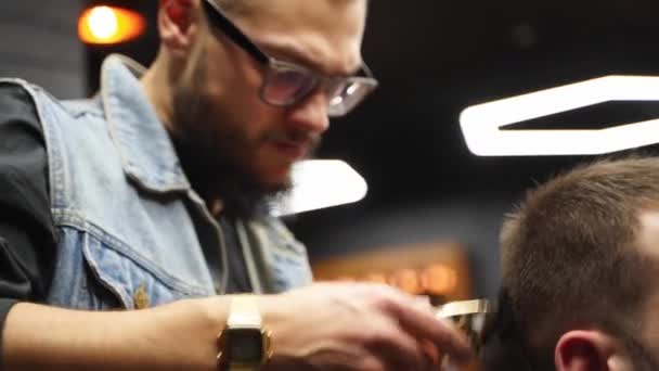 Trendy barber cuts bearded mans hair with a clipper in barbershop. Mens hairstyling and hair cutting in salon. Grooming the hair with trimmer. Hairdresser doing haircut in retro hair salon. — Stock Video